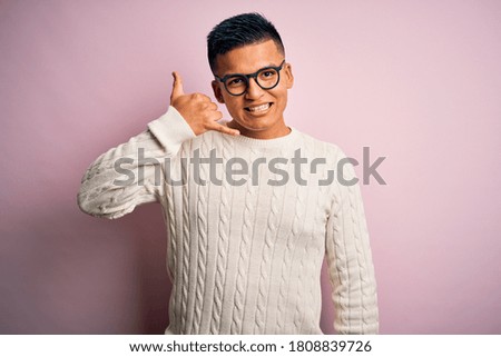 Young handsome latin man wearing white casual sweater and glasses over pink background smiling doing phone gesture with hand and fingers like talking on the telephone. Communicating concepts.
