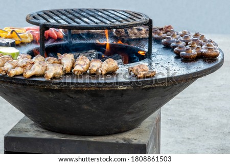 Large round metal charcoal grill with chicken legs, mushrooms and vegetable slices. Selective focus.