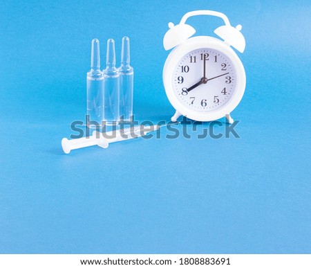 White alarm clock, medical ampoules, syringe on a blue background with copy space. The concept of vaccination against infection and viruses