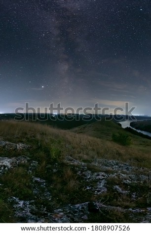 Milk Way night sky over hills and Dniester river. Night nature landscape. City lights on background. Ukraine. Vertical photo