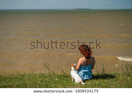 Rear view of a sitting woman on the river bank with a dog. Girls spend weekends in nature with a pet.