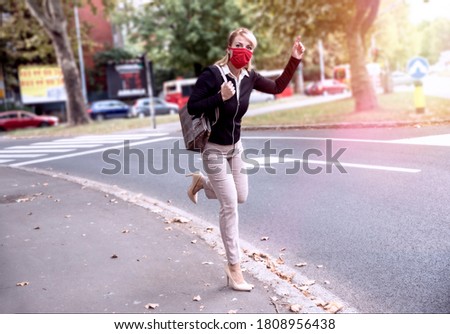 Woman with red protective cloth mask on face waving to a taxi on City street. Businesswoman late on job during Corona virus pandemic. Full length of Female running to work Royalty-Free Stock Photo #1808956438