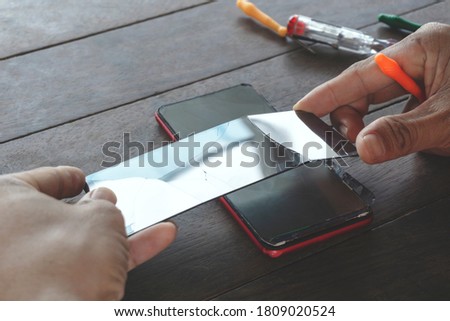 A man technician holds a black smartphone screen and mini tools cracked or broken from a drop for repair or cell phone replacement, with a red phone body on a wooden table with a screwdriver.