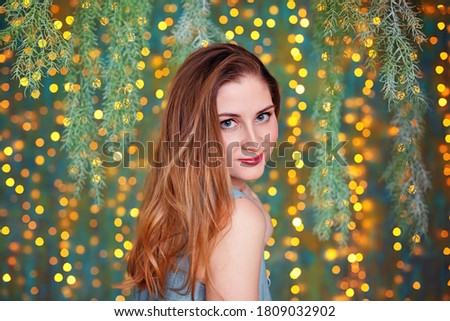 A young beautiful woman in an evening dress with loose red hair stands in the room against the background of lights garlands