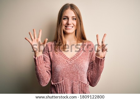 Young beautiful redhead woman wearing pink casual sweater over isolated white background showing and pointing up with fingers number seven while smiling confident and happy.
