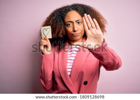 Young african american woman with afro hair holding paper with heart over pink background with open hand doing stop sign with serious and confident expression, defense gesture