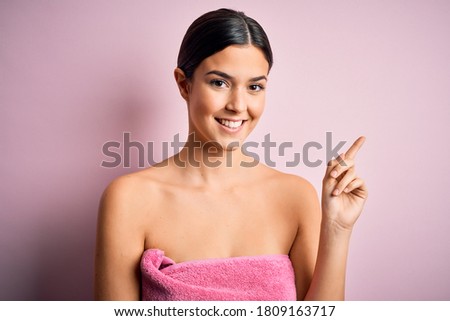 Young beautiful girl wearing towel shower after bath standing over isolated pink background with a big smile on face, pointing with hand and finger to the side looking at the camera.