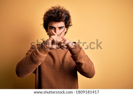 Young handsome man wearing casual shirt and sweater over isolated yellow background Rejection expression crossing fingers doing negative sign
