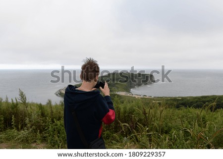 The sea, the hills, the peninsula, the boy stands with his back and takes a photo with his phone.