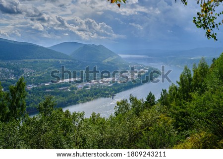 View from the mountain to a wide river with small houses on the Bank. Green forest and dark blue sky before a thunderstorm