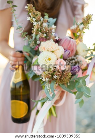 Woman dressed in pastel dress holding bottle of shampagne and messy bouquet of exotic flowers, roses and dahlias, shallow depth of field emotional details