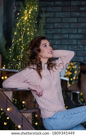 Young woman which sits on a wooden staircase among Christmas trees and garlands with lights. A girl smiles, dressed in a soft pink knitted sweater, blue jeans, with curly, long hair. Close-up portrait