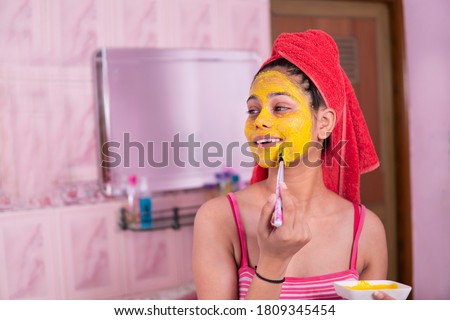 Skin care - Beautiful girl applying Gram flour turmeric yellow face mask on face through brush. She is wearing red towel on head. Royalty-Free Stock Photo #1809345454