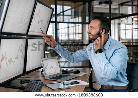 Bearded man trader wearing headset with microphone sitting at desk at office in front of monitors with data candle chart doing online trading training talking to employee explaining supply and demand Royalty-Free Stock Photo #1809388600