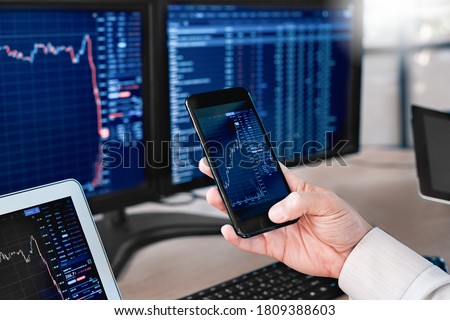 Man trader sitting at desk at office in front of monitors with stocks data holding smartphone browsing application monitoring price changes on candle bar online using digitla devices close-up Royalty-Free Stock Photo #1809388603