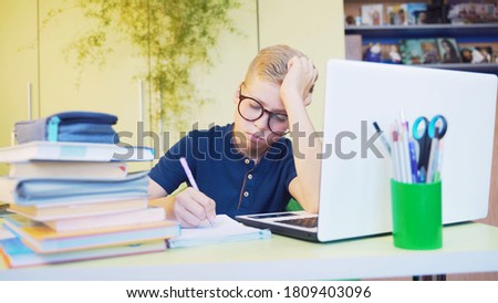 Cute tiring teenage boy sitting at the table and writing at notebook during distance learning at home, with eyeglasses. Boring child, schoolboy during online studing with books and white laptop.