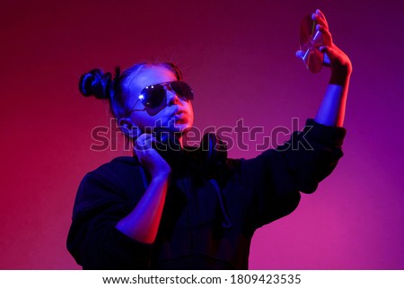 fashionable teenager girl in glasses and with headphones looks at a cd disk on a pink background and in blue-red disco lighting