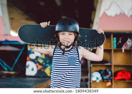 Portrait of handsome caucasian boy athlete skateboarder in protective helmet with skateboard in hands looking at camera on background of skate park. A child and an active hobby, sports and health.