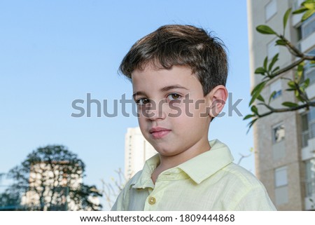 Brazilian child, 7 years old, serious, on a sunny afternoon. In the background the blue sky and a building.