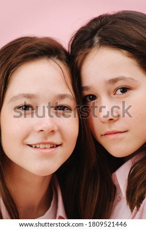 Beautiful little twin sisters pressing heads to get into image frame. Over pink background, studio shot. Both looking at the camera, one smiles openly, her sister has closed smile.