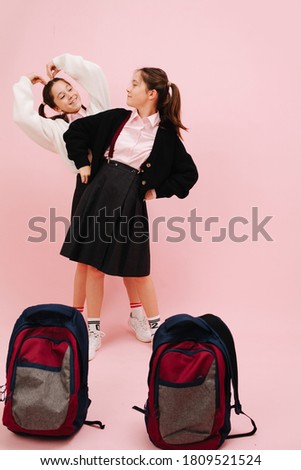 Beautiful little twin schoolgirls with twintails happily posing in uniform. Low angle. Over pink background, studio shot. One standing behind the other, looking at each other. Backpacks on the ground