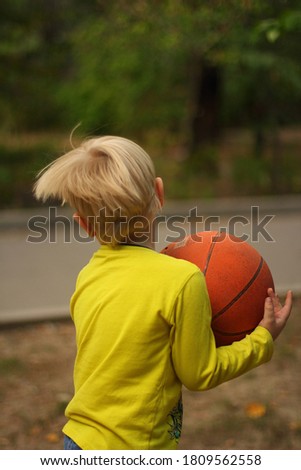 A little boy with blond hair in a lemon-colored jacket holds a basketball ball in his hands