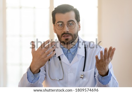 Head shot portrait young doctor wearing glasses and white uniform with stethoscope speaking, consulting patient online, looking at camera, making video call, sitting at table in office