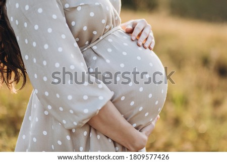 A pregnant woman in a dress holds her hands on her stomach on a nature background. Pregnancy, motherhood, preparation and expectation concept. Close-up, Beautiful gentle mood photo 
