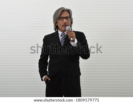 Confident elderly asian businessman in black suit and eyeglasses speaking with microphone standing over white wall background, Business presentation and seminar concept