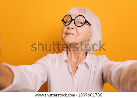 Portrait of elderly woman with short white hair wearing white glasses and light blouse holding camera and taking selfie. Old Woman isolated over orange background Royalty-Free Stock Photo #1809601786