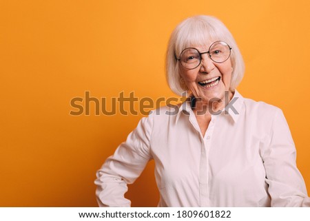 Image of happy old woman with light short hair wearing white blouse holding her arms on hips and smiling. Woman isolated over bright orange background Royalty-Free Stock Photo #1809601822