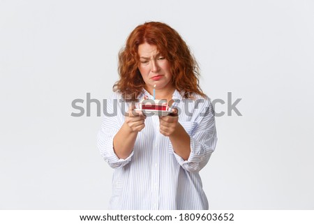 Celebration, holidays and emotions concept. Uneasy and sad redhead middle-aged woman hate her birthday, looking upset at b-day cake with lit candle, feeling mid-life crisis, white background