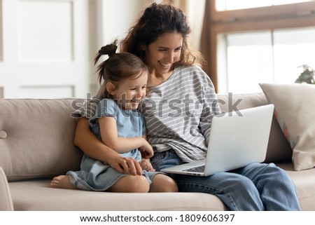 Happy mother with little daughter have fun online with new cool app, family watching cartoons, play educational game on laptop sit on sofa. Enjoy video call talk, develop child use modern tech concept