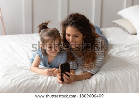 Mother lying on bed with little daughter curious cutie child involved in video call by smart phone with relatives enjoy distant talk. Modem tech and kids, parental control and fun on internet concept