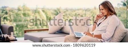 Attractive woman 40 years old in a white shirt sitting on a gray sofa working on a laptop on the terrace overlooking the green jungle on a bright sunny day.