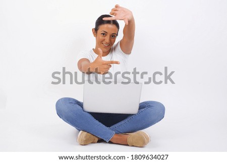 Portrait of young positive female using computer with cheerful expression, dressed in casual light blue sweater, has good mood, gestures finger frame actively at camera.