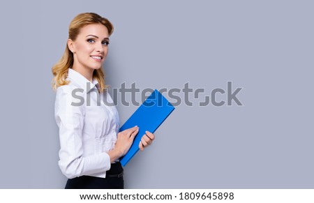 Happy smiling businesswoman holding blue folder, with copy space area for some slogan or text. Confident blond woman in white shirt, isolated over grey color background. Success in business concept. 