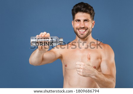 Smiling young bearded fitness sporty strong man bare-chested muscular sportsman posing isolated on blue background. Workout sport motivation lifestyle concept. Hold bottle of water, showing thumb up