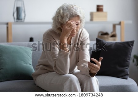 Unhappy stressed mature middle aged woman looking at phone screen, sitting on couch in living room, upset frustrated senior female reading bad news in message or social network, problem with device Royalty-Free Stock Photo #1809734248