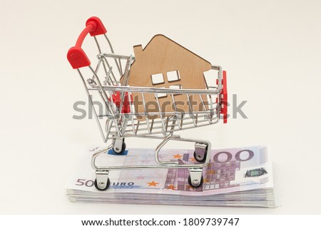a small wooden house lies in a cart from a supermarket, the cart is on a wad of money, euros. Concept - purchase, acquisition, real estate, affordable housing, mortgage. Horizontal photo, close-up