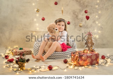 Christmas studio shoot of a cute baby girl that is hugging a teddy bear. Festive, beige background with gifts, Christmas balls and lights