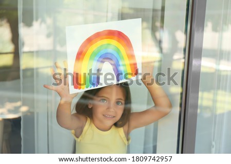 Little girl with picture of rainbow near window, view from outdoors. Stay at home concept