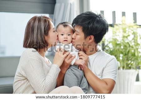 Happy young Vietnamese mother and daughter kissing baby girl on both cheeks