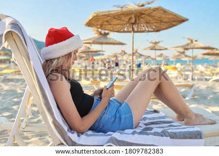 Young woman with smartphone in hat of Santa Claus resting sitting on beach in sun lounger. Christmas and New Year holidays, vacations, travel, tourism concept