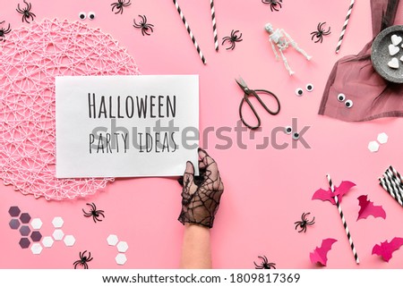 Halloween party ideas text on white page held in hand. Flat lay with scissors and decorations on pink paper background. Hexagon confetti, paper drink straws, bats, sugar hearts and spiders.