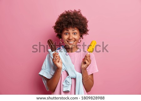 Summer time concept. Glad positive woman with Afro hairstyle holds tasty frozen ice cream, enjoys eating delicious cold dessert, dressed casually, poses against pink background, feels happy.