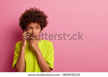Photo of serious dark skinned woman holds chin, has telephone conversation, dressed in casual bright clothes, looks aside, poses against pink background, blank copy space for your advertising content