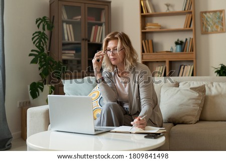 Serious mature blond businesswoman in casualwear sitting on couch by small table in front of laptop while networking in living-room