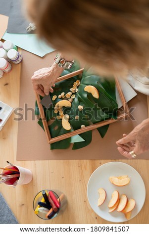 Top view of hand of mature woman making picture composition in wooden frame while putting apple slices on green leaf of domestic palm