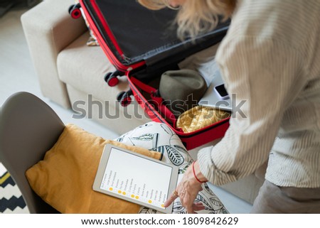 Mature woman pointing at touchpad display with list of things to take for journey while putting clothes and other stuff into suitcase before trip
